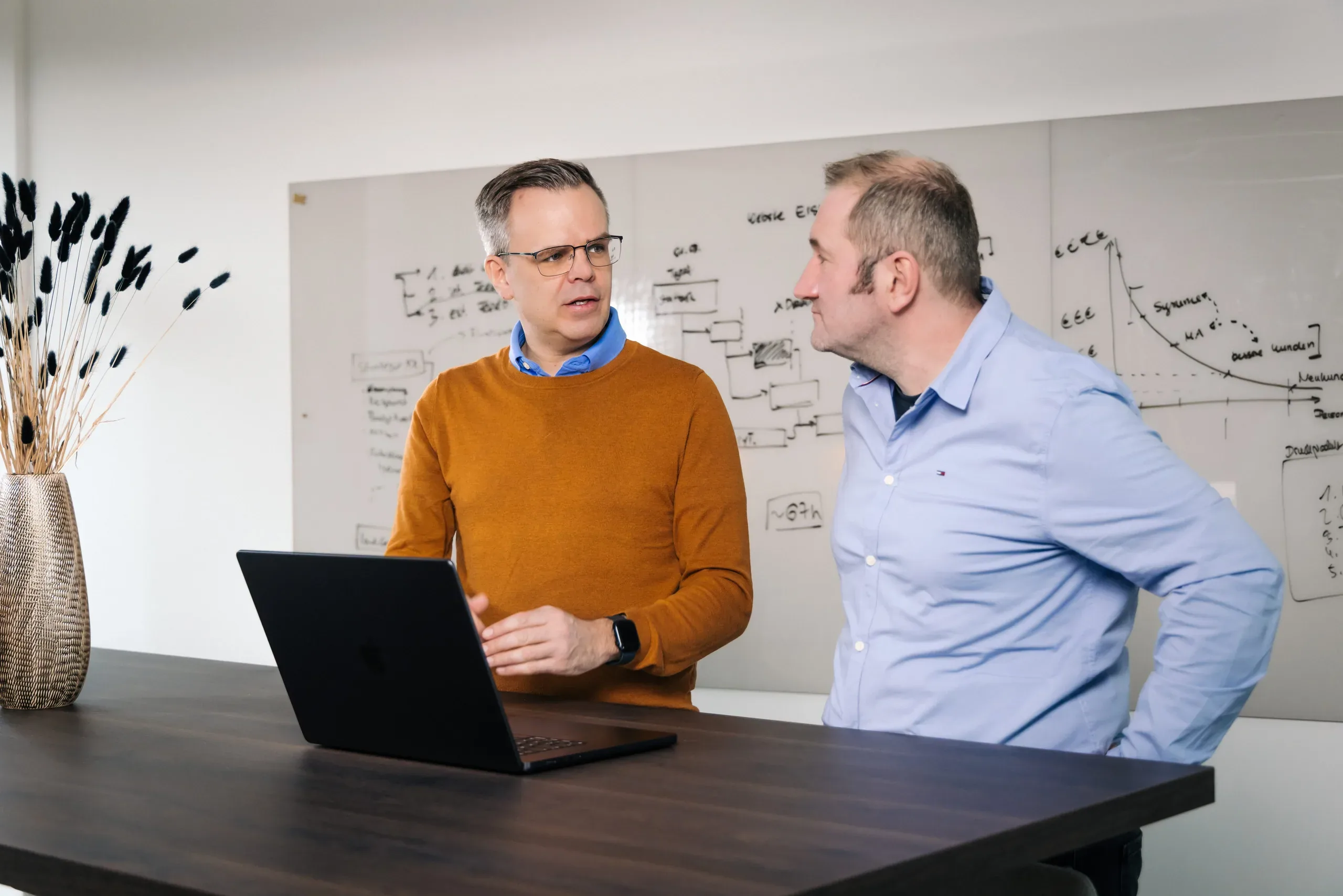 Zwei Kollegen besprechen Kursdaten am Laptop in der Küchenecke